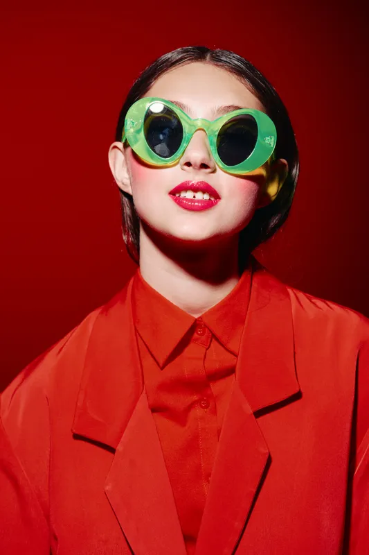 Stylish woman with bright red lipstick wearing oversized green sunglasses and a red outfit against a vibrant red background. Concept of beauty, fashion, creative style, and modern trends.