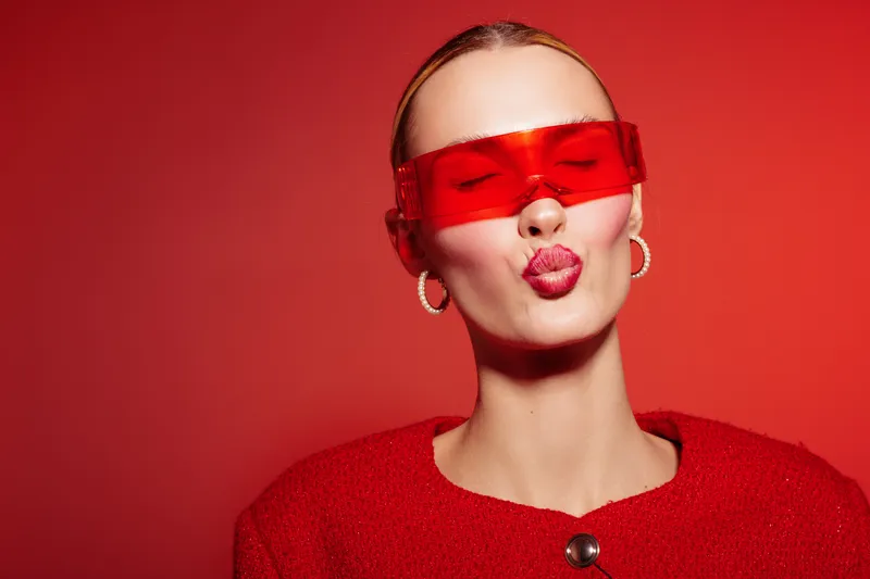 Young woman in red sweater and red translucent glasses blows a kiss on red background. Fashion portrait with bold makeup and stylish accessories. Concept of beauty and modern style.