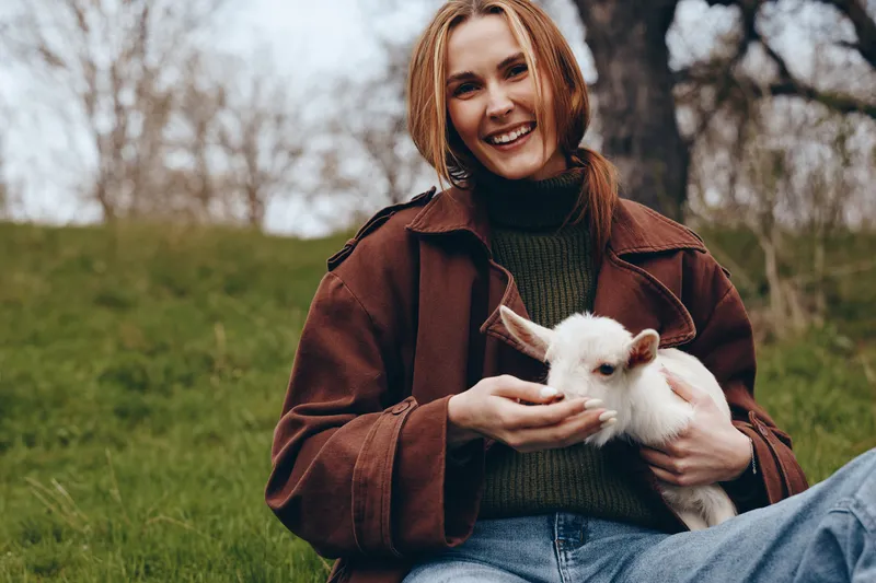 Joyful woman outdoors with a white rabbit in her arms, wearing a brown coat and green sweater, a cozy lifestyle moment in a park, celebrating companionship with a gentle animal