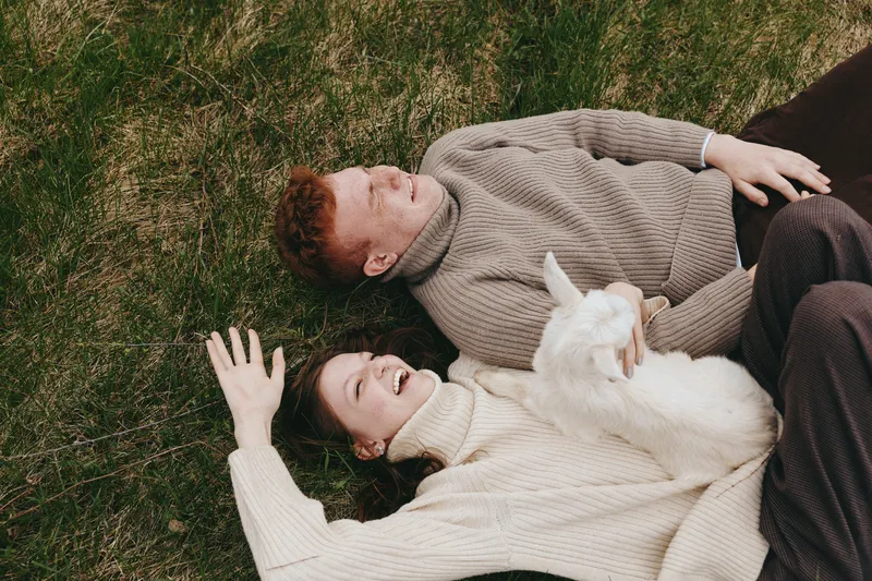 Cheerful couple outdoor, relaxing on grass with a playful goat kid, soft sunlight, cozy knitwear, natural tones, calm interaction, carefree mood, nature backdrop, lifestyle moment