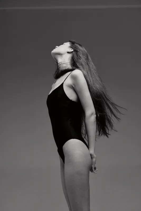 Black and white photo of a woman in a black swimsuit looking upwards outdoors with long hair flowing and calm expression