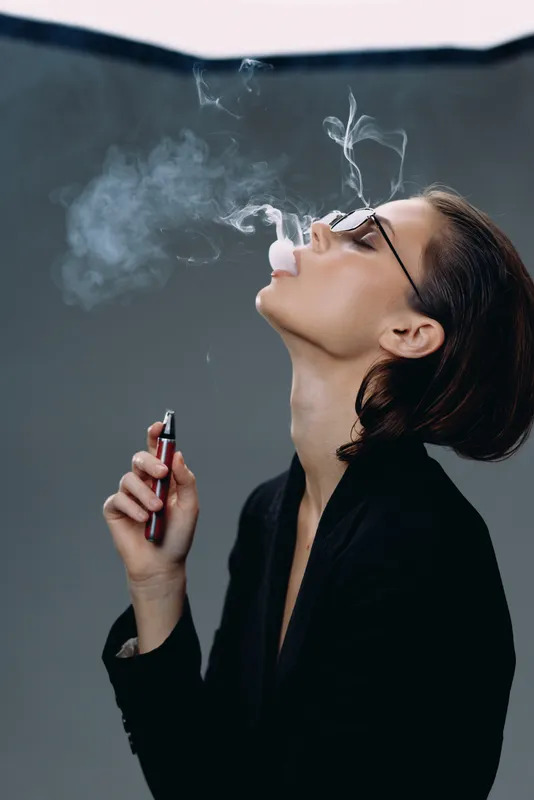 Young man vaping and blowing smoke rings with e-cigarette wearing glasses and black jacket