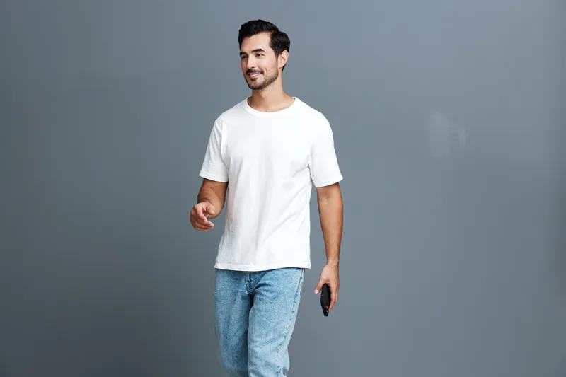 Happy smiling young man in casual white t-shirt walking forward while holding a smartphone, with a neutral gray background. Lifestyle and modernity concept.