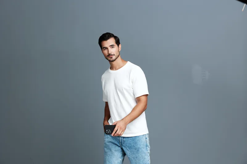 Young smiling man in a casual white t-shirt and denim jeans poses confidently, holding a mobile phone, against a muted gray background. Lifestyle concept.
