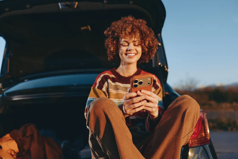 Woman lifestyle with curly hair, smiling, holding smartphone while sitting on car trunk outdoors at sunset, casual roadtrip mood, warm light, relaxed selfie portrait, evening sky