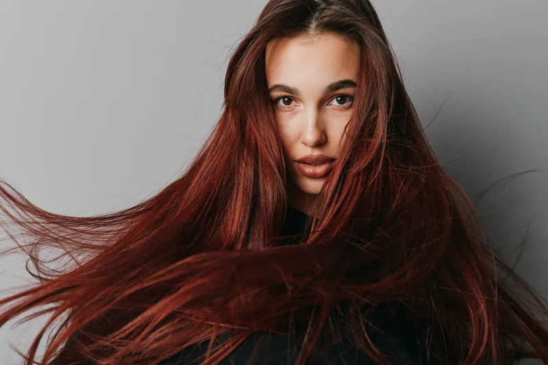 Woman with long red hair blowing in wind looking at camera with serious expression on gray background. Portrait of female model with natural makeup and loose hair movement.