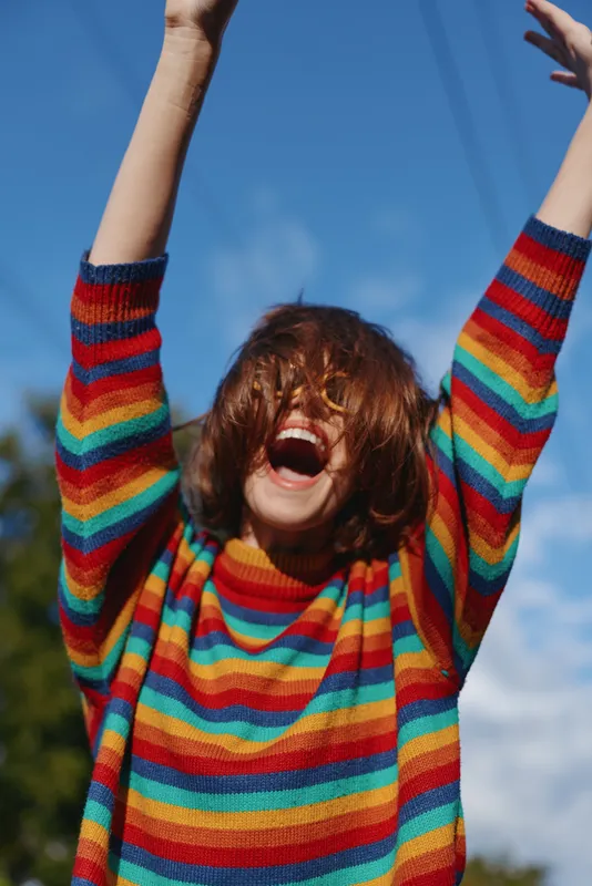 Woman joyful in her twenties wearing a rainbow sweater and striped knit, laughing with arms raised outdoors under blue sky. Casual outfit, carefree lifestyle portrait for fashion, travel and street
