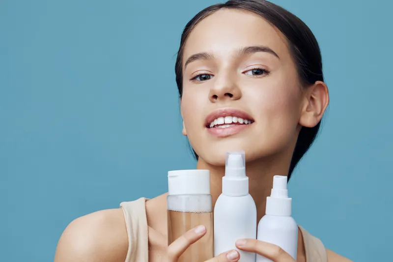 Young woman showcasing skincare products, smiling confidently against a solid blue background, highlighting natural beauty and self care