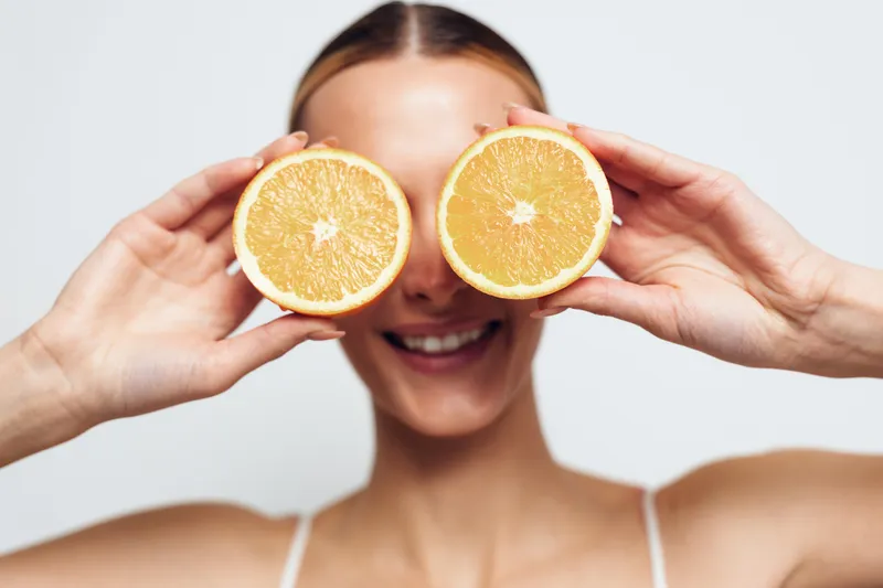 A smiling woman holds two halves of an orange in front of her eyes against a plain white background. The fresh fruit and cheerful expression convey health and vitality.