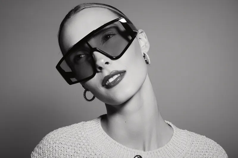 Close-up of a fair skinned Caucasian woman wearing oversized geometric sunglasses and hoop earrings posing in studio lighting. Fashion concept with minimalist style and elegance.