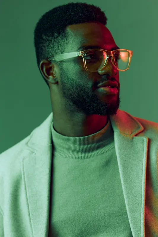 Stylish portrait of a young Black man wearing glasses and a cozy sweater, exuding confidence and creativity against a vibrant green backdrop