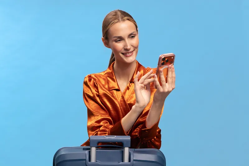 Smiling woman in orange blouse uses smartphone while sitting behind suitcase against blue background. Concept for travel, communication, technology, and leisure with positive mood.