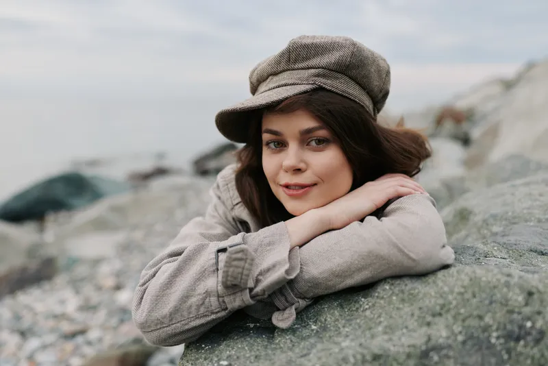 Young woman in beige coat and hat leans on rock by the beach. She looks relaxed and happy with soft natural light and calm sea in the background.
