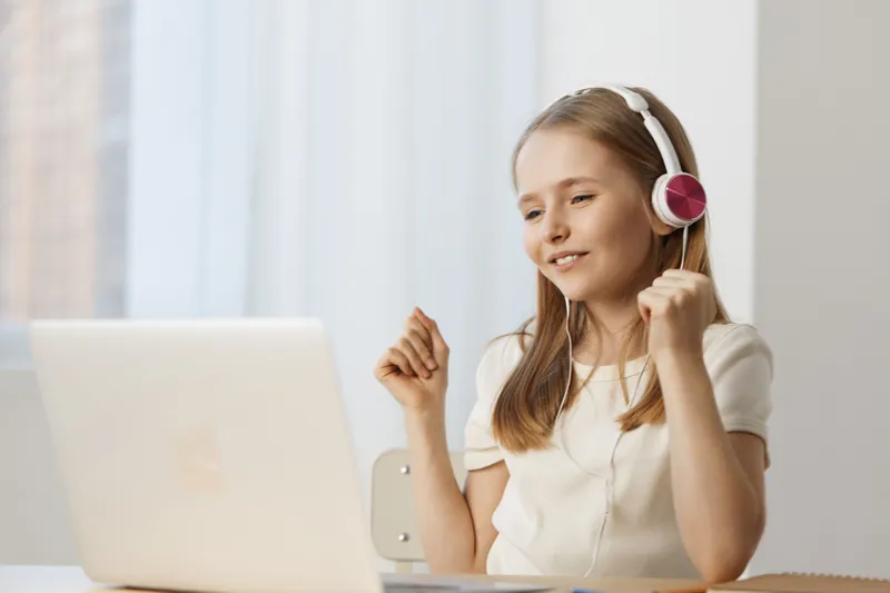 Young girl enjoying music while using a laptop, wearing headphones, expressing joy and engagement in a bright and airy room