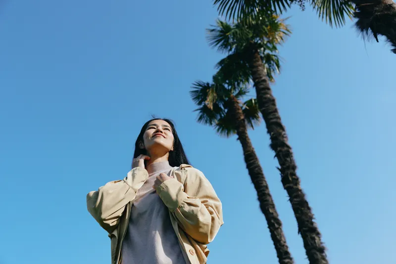Woman outdoor sky palm trees looking up casual fashion confidence under bright daylight, wearing light jacket and sweater, serene mood and warm sunlight in open sky