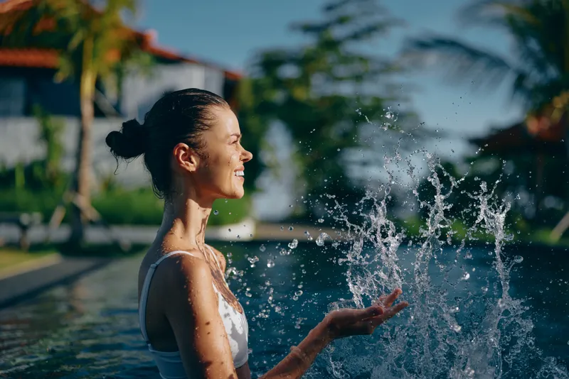 A joyful woman in a white swimsuit enjoys a sunny poolside moment, water droplets arc through the air as she smiles, capturing a carefree lifestyle scene by the pool.