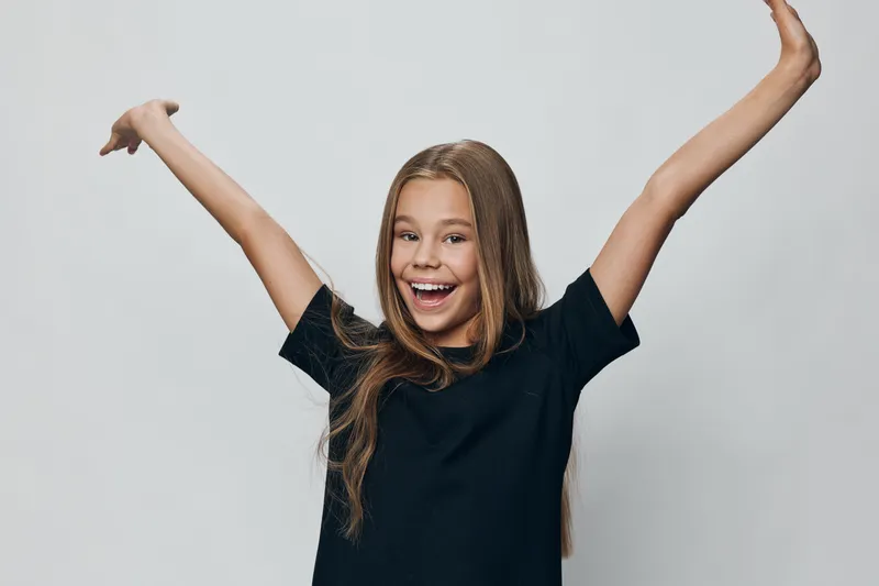 Happy girl with long hair wearing black shirt smiling and raising arms up isolated on grey background. Child, emotion, joy and expression for childhood lifestyle and positive energy.