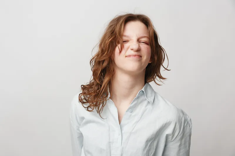 Young woman with curly hair wearing white shirt makes funny face with closed eyes and scrunched nose. Isolated on white background. Concept of humor, emotion, and playful expression.