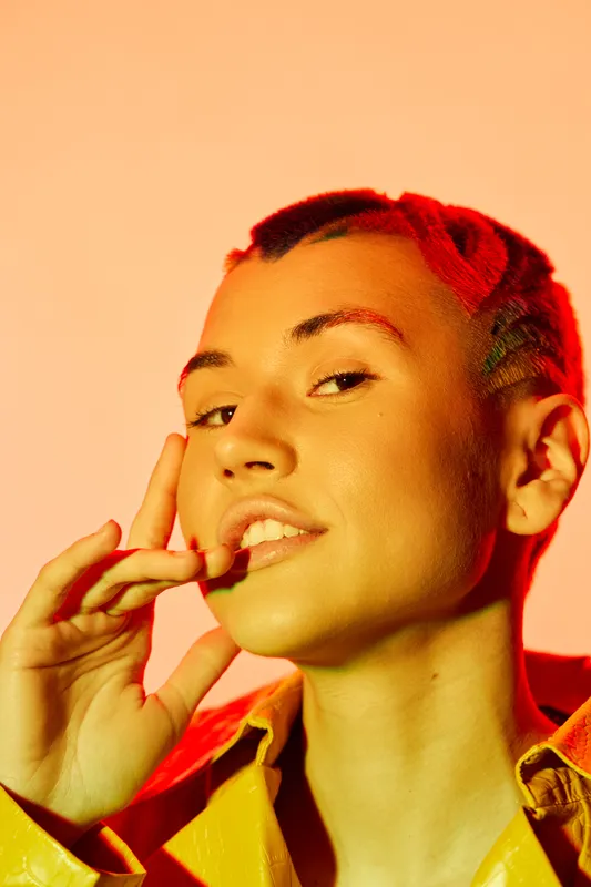 Close-up portrait of woman with braided hair and yellow jacket posing confidently against warm orange background, showcasing bold attitude and modern style.
