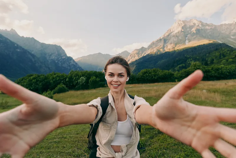 Cheerful woman outdoors, smiling toward the camera, arms extended, sunlit meadow with trees, distant mountains, casual outfit, natural colors, lifestyle moment