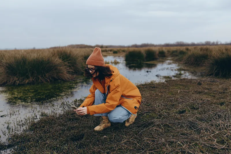 Woman lifestyle outdoors by riverbank, kneeling in warm orange jacket and hat, smiling and listening to nature sounds, soft cloudy light, calm mood, grasses and reeds background
