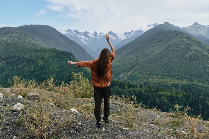 Person standing on a rocky overlook with arms raised, gazing at mountains and a pine forest valley, conveying triumph, exploration, and a sense of freedom