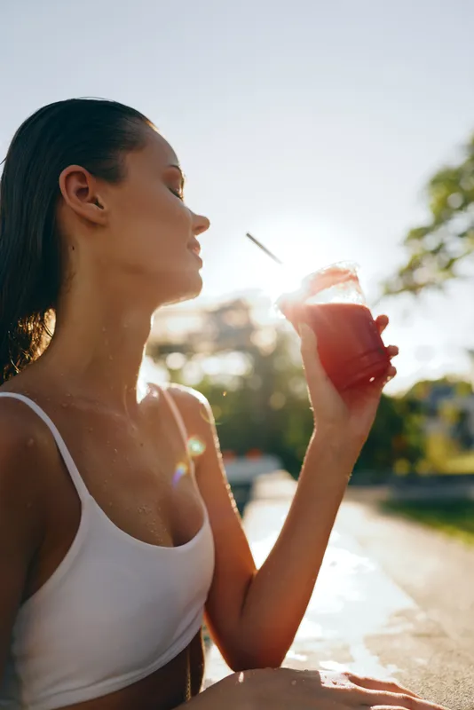 Woman in a white tank top enjoys a sunlit outdoors moment, sipping a drink by the poolside, conveying a casual lifestyle scene of summer relaxation and serenity, capturing warm glow and carefree mood.