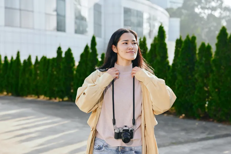 Woman with camera hanging from neck outdoors, wearing beige jacket and light sweater, standing beside green hedges near a modern building, ready for a photography shoot