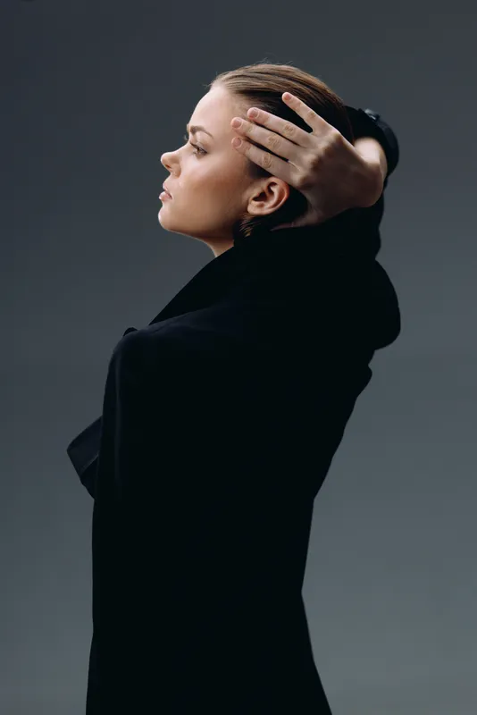 Profile of a thoughtful woman in black clothing with hands on head against dark background
