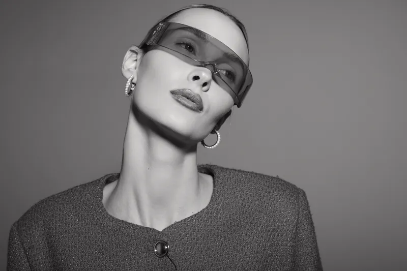 Stylish white woman with pale skin wearing futuristic sunglasses and textured jacket looks confident in black and white studio portrait.