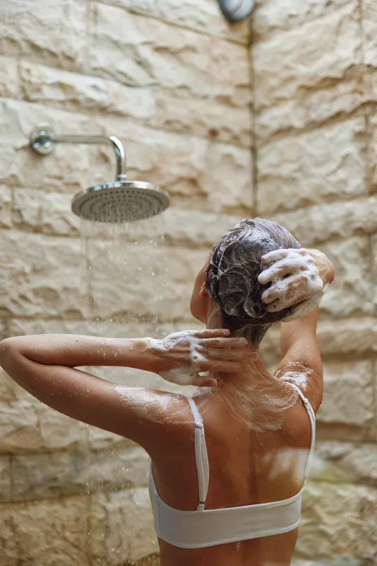 woman washing hair under modern showerhead with stone tile background, soap foam on skin, natural skincare, personal hygiene, wellness routine