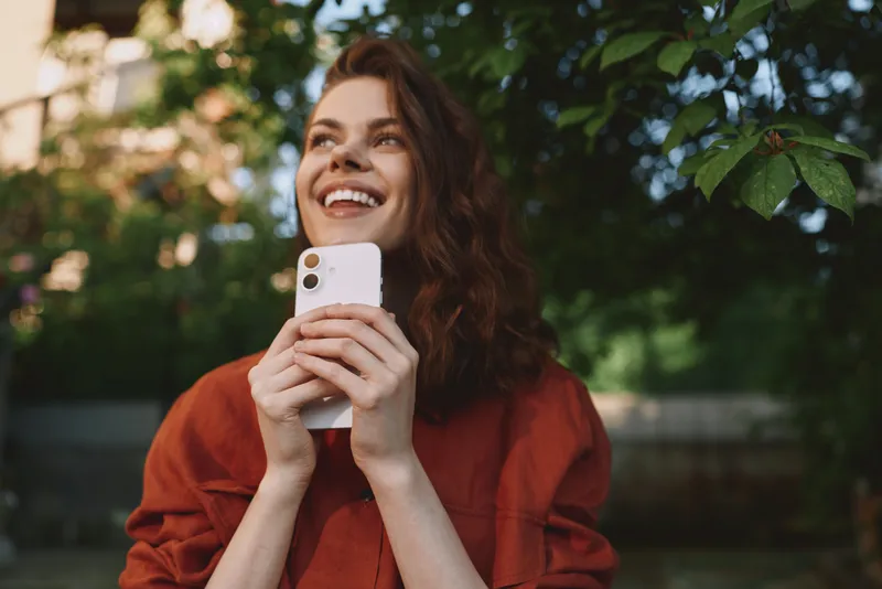 Woman lifestyle portrait outdoors, smiling while holding smartphone, natural light, greenery background, casual red top, candid moment of connection and joy