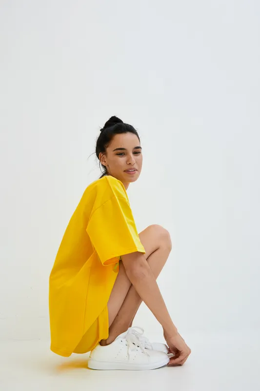 Young woman wearing yellow oversized t-shirt and white sneakers, sitting crouched with a relaxed expression against a minimal white background.