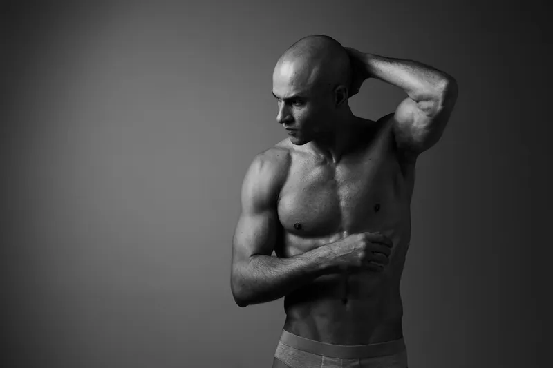 Black and white portrait of a man showing his strong physique and thoughtful expression, highlighting his muscular detail and emotional depth.