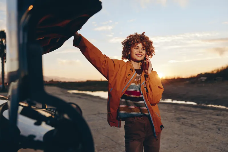 Woman lifestyle outdoor at sunset near car, curly hair woman in jacket stands by open door, smiling and raising arm, warm golden light, travel mood