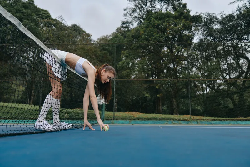 Young female athlete in tennis attire preparing to serve on a blue court surrounded by green trees, showcasing focus and determination in her sport