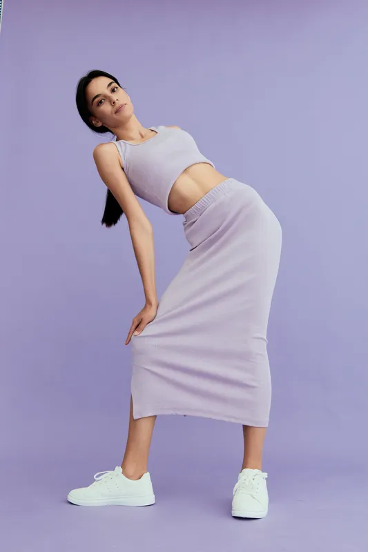 Slim woman wearing a sleeveless crop top and matching long skirt leans gracefully against a lilac background, showcasing modern casual style with white sneakers.