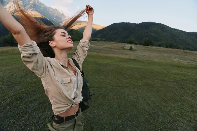 Vibrant woman lifestyle outdoors, arms raised in joyful moment, sunlit meadow with distant mountains, relaxed posture, natural colors, fresh breeze, clear sky bright sunlight glow