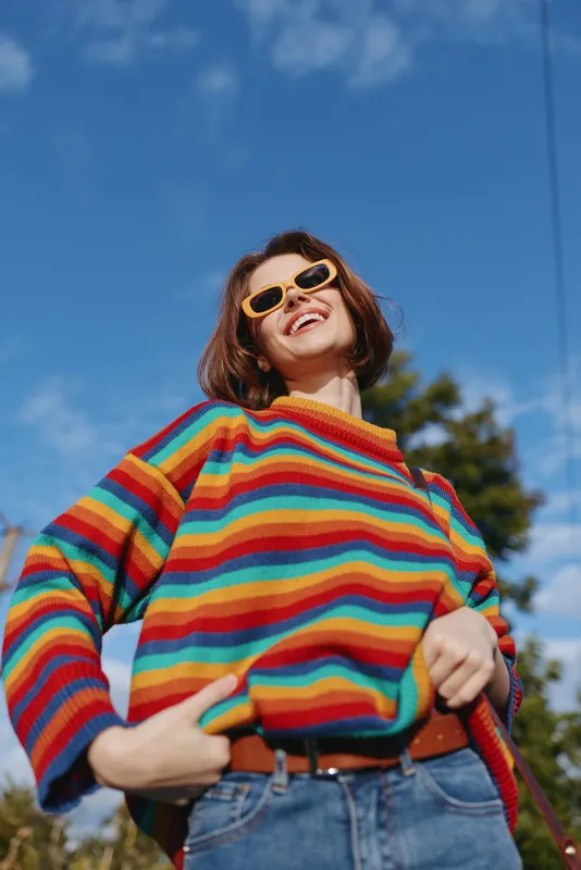 woman smiling in striped sweater and yellow sunglasses, young adult with short brown hair, casual outfit with jeans, hands on belt, laughing outdoors under blue sky for lifestyle fashion