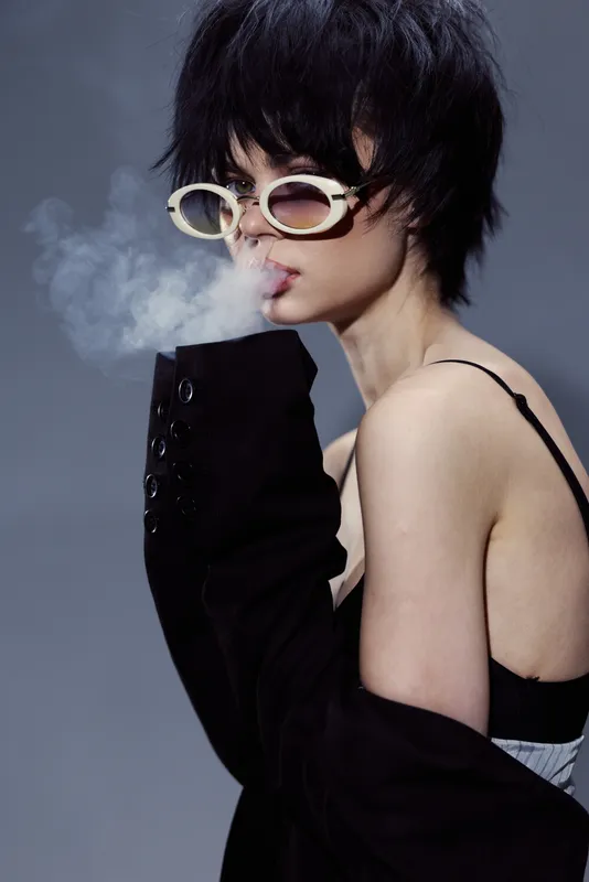 Stylish woman with short black hair wearing sunglasses and blowing smoke in a black outfit and exposed shoulder