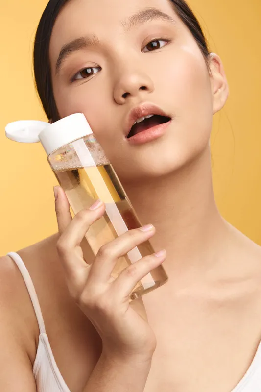 Asian woman holds a clear bottle of skincare near her face, looking at the camera with a soft expression. The warm backdrop highlights healthy skin and a calm, modern beauty routine.