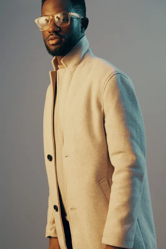 Stylish African male model wearing a light coat and glasses, exuding confidence against a soft gradient background ideal for fashion photography