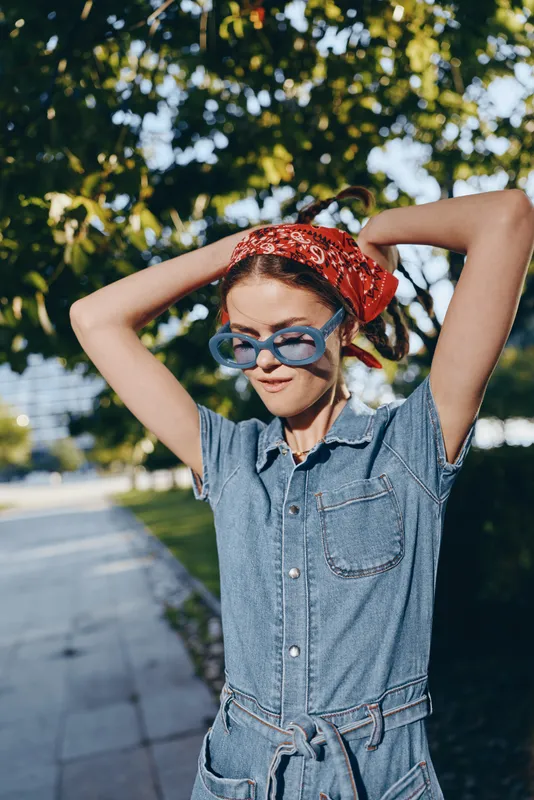 Lifestyle woman in modern boho-western denim street style wearing blue sunglasses and red bandana, captured with warm film color outdoors on sunny day.