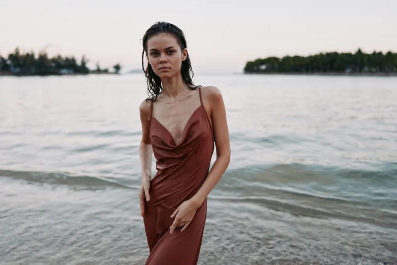 A solitary woman stands at the shoreline in a deep red satin dress, facing the camera as gentle waves roll in and distant trees frame a serene seascape.