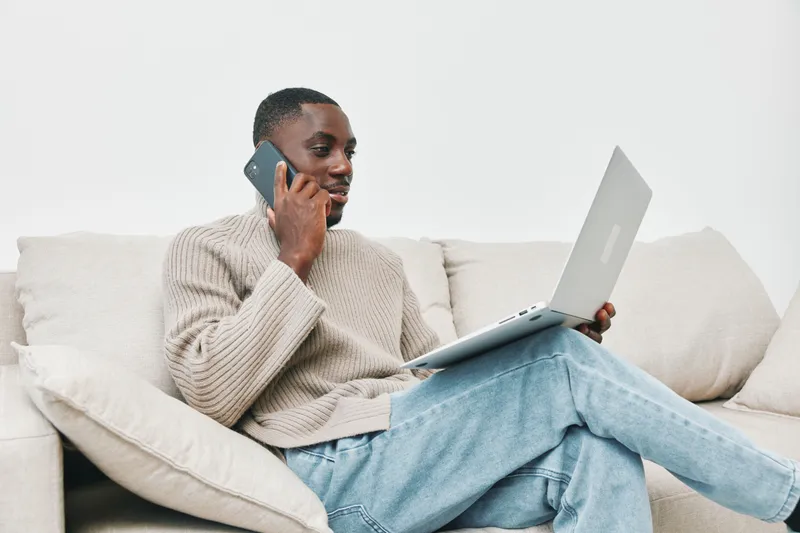 Young Black man using smartphone and laptop for remote work, wearing casual beige sweater and jeans, relaxed atmosphere, modern interior with soft tones
