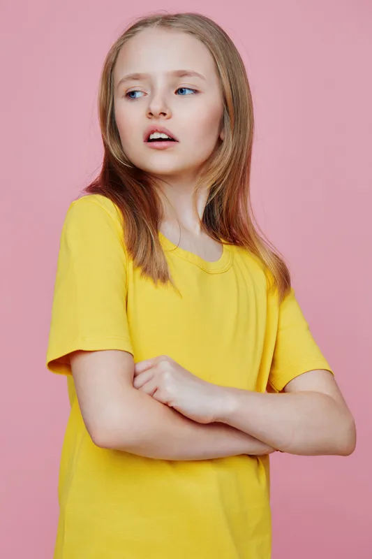 Young girl with long hair in a yellow shirt posing confidently against a pink background, expressing a sense of individuality and youthfulness