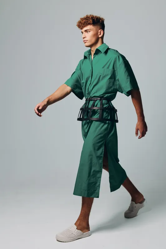 Young man walking in green jumpsuit with short sleeves and wide legs, wearing white slip-on shoes and a black belt accessory, posing in studio with neutral background.