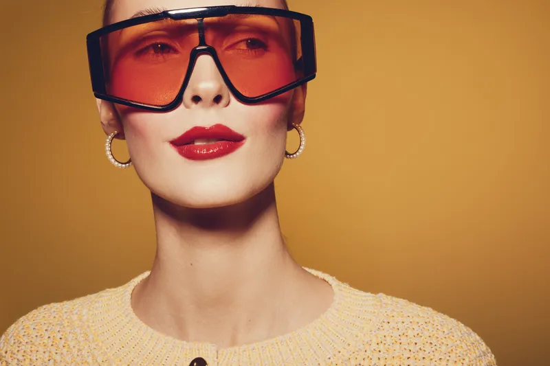 Stylish woman with red lipstick wearing oversized orange sunglasses and hoop earrings poses against yellow background. Fashion portrait with retro vibe and confident expression.