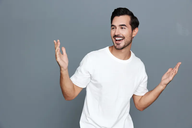 Smiling happy cheerful young man wearing a white t-shirt gestures with open hands, expressing joy and excitement against a grey background. Positive lifestyle concept.