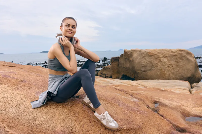 Young athletic woman sitting on rocks by the sea, wearing fitness clothing, smiling confidently, showcasing a healthy lifestyle and tranquility